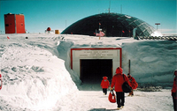People wearing cold weather gear walk towards a square, tunnel-like entrance surrounded by snow. The dome at the South Pole Station is in the background.