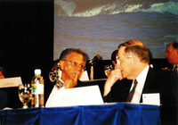 Warren, left, and Neal, right are seated next to each other and looking towards each other.