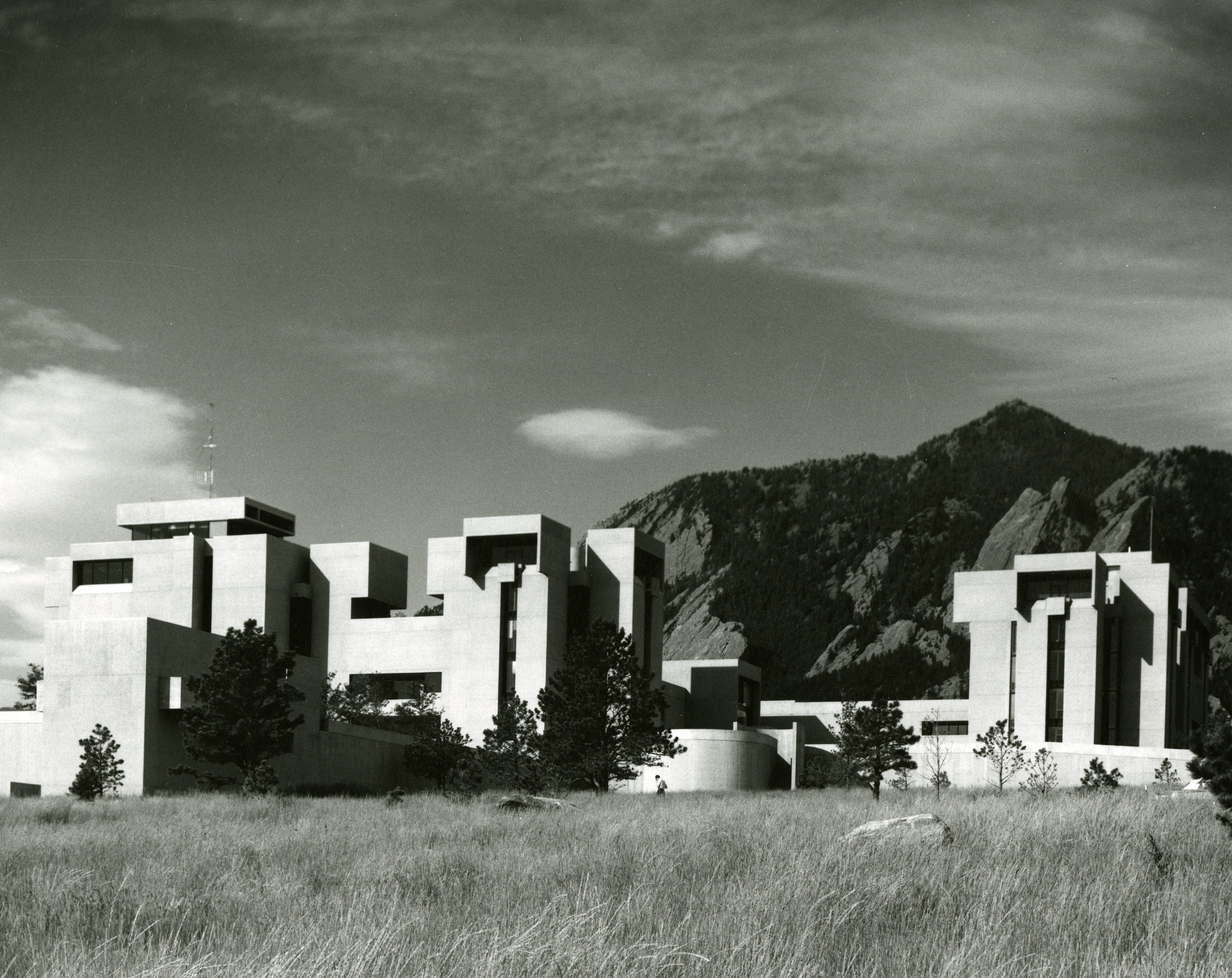 Photo of the Mesa Lab taken with the Flatirons in the background, wispy clouds overhead, and wild grasses in the foreground. 