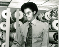 Warren Washington stands in front of rows of magnetic data tapes. He is looking down and to the right.