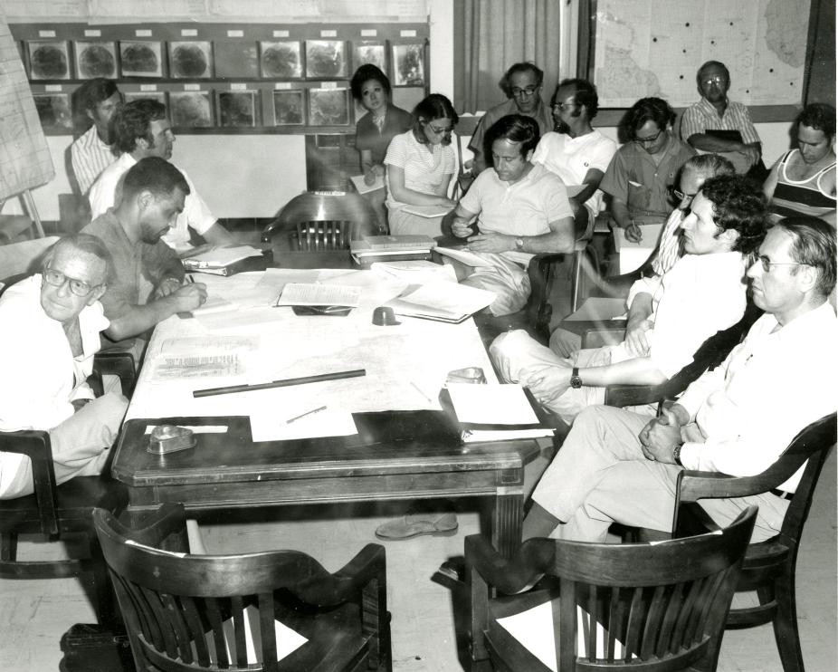 A group of people sit around a table that is covered in maps and papers. There are maps and satellite photos on the wall behind the people. Joachim Kuettner is on the far left facing the camera.