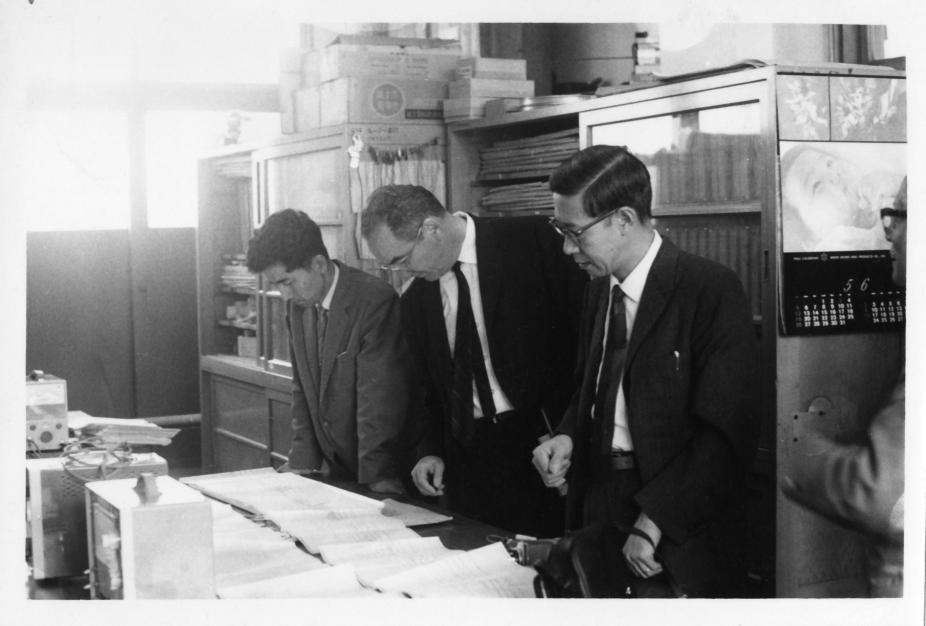 In a room crowded with cabinets, Walter Orr Roberts, I.M. Pei, and two others look at papers that are spread across a table in the middle of the room.