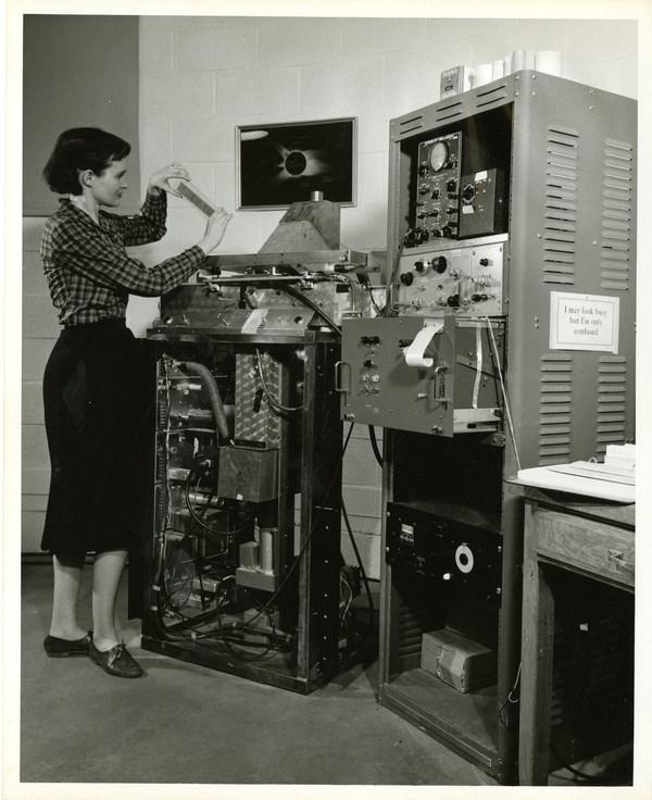 A woman stands in front of an elbow-high piece of equipment holding a strip of photographic negatives.