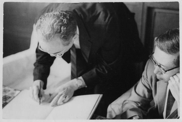 Walter Orr Roberts writes in a book. I.M. Pei, sitting to the right of Roberts, observes.