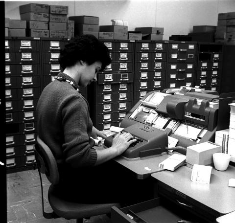 Photograph of a woman entering data into an IBM 26 punch card printer. She is faced away from the camera and in a room lined with filing cabinets. The photo was taken in 1966 in the NCAR Computer Room.