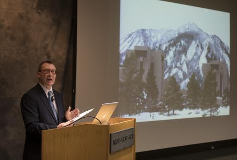 Stuart Leslie stands in front of a podium giving a presentation. A projected image of the Mesa Lab is behind him on the stage.