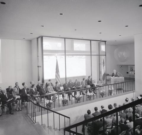 Mesa Lab lobby seen from above. A couple of rows of people are seated on the mezzanine next to a podium where a man is standing and speaking. An audience is seated below facing the mezzanine.
