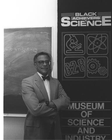 Warren Washington, wearing a suit and bowtie, stands in front of a banner that says "Black Achievers in Science: Museum of Science and Industry".