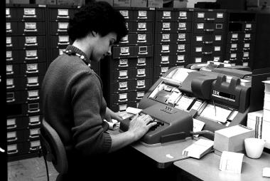 Photograph of a woman entering data into an IBM 26 punch card printer. She is faced away from the camera and in a room lined with filing cabinets. The photo was taken in 1966 in the NCAR Computer Room.