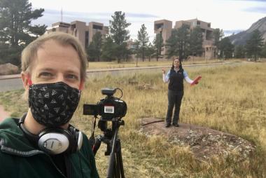 Dan Zietlow, wearing a mask, stands at a camera. Lorena Medina Luna is standing at a distance in front of the camera. 